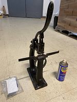 Photo of the full pamphlet stapler next to a WD-40 spray can and pack of staples, showing entire machine with handle and base.