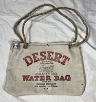 Front view of canvas Desert Brand camping water bag with red and black printed text, rope handles and metal screw cap spout.