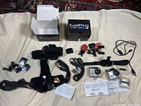 Overview of lot showing two GoPro cameras in waterproof cases, mounting straps, various cables, clips, and box packaging.