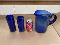 Hand made blue glass pitcher and two glasses shown with a coke can for scale