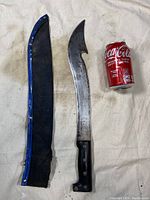 Full view of the machete with black handle beside its blue and black sheath and a standard 12 oz Coca-Cola can for scale.