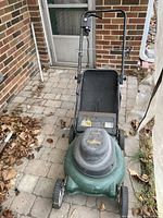 Outdoor photo of green Yardworks electric lawn mower with grass catcher and upright handle, placed on a patio with fallen leaves around.