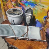 Image showing the rice cooker with glass lid, the warming tray with power cord, and the can opener, displayed on a wooden surface with colorful backdrop.