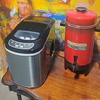 Photo shows side-by-side modern ice maker machine and vintage red coffee maker on wooden table.