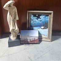 Three art pieces together: one statue and two paintings, displayed on a grey surface against a brown background.