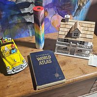 Photo showing the four main items on a wooden surface: yellow ceramic car figurine with cartoon characters, colorful kaleidoscope, Rand McNally World Atlas book, and miniature wooden house model.