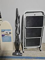 Ironing board folded with beige cover, Bissell vacuum upright, and folded white step ladder.