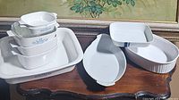 Stacked white CorningWare baking dishes with blue floral pattern and several white ceramic baking pans of assorted shapes on a wooden surface with a floral backdrop.
