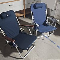 Photo shows two blue fabric beach chairs with metal frames, one with wood veneer armrests and the other with plastic armrests, both with back pillows and cup holders.
