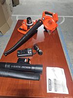 Full view of cordless leaf blower with blower tubes attached and detached, battery pack, charger, and manual laid out on a wooden table.