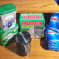Photo showing two green containers of Cascade dishwasher pods, one Platinum with 24 pods and one Original with 105 pods, along with a box of Husky outdoor drawstring trash bags and two packs of Windex wipes on wooden surface.
