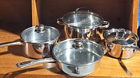 Four stainless steel cookware pans with glass lids arranged on wooden shelf, varying in size and design, showing visible wear