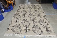 Full view of tan rug with repeating black and grey floral patterns, showing fringe edges.