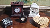Front view showing all five items on a wooden surface beneath a floral wallpaper background, including the three clocks and two decorative boxes.