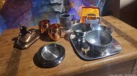 Overview of all metal pieces including cups, bowls, plates, tray, and decorative boot sculpture on wooden table with colorful background