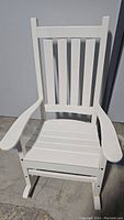 Front view of the white wooden outdoor rocking chair showing vertical slats on the back and seat with wide armrests and rocker legs.
