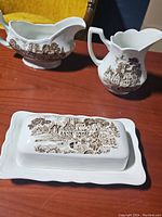 Gravy boat, butter dish with lid and base, and pitcher displayed on wooden surface with yellow cushion in background