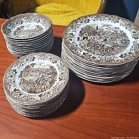 Stack of dinner plates, salad plates, and small bowls showing brown transferware pattern with scenic village and floral border design.