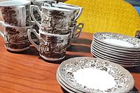 Stack of 6 tea cups with brown pastoral design and two stacks of saucers with floral border on wooden table