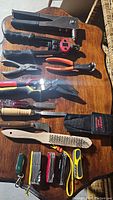Various hand tools including wire cutters, pliers, a stapler, chisels, a wire brush, and multi-tool knives on a wooden surface