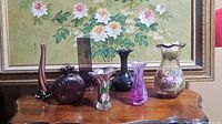 Photo showing all seven purple glass vases arranged on a wooden surface with a floral painting background.