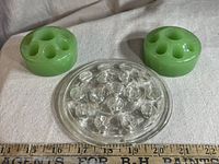 Three vintage flower frogs on fabric with measuring tape: one clear crystal with 16 holes and two green jadeite with 7 holes each, showing cracking in jadeite.