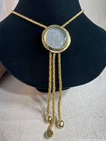 Front view of the gold tone necklace displayed on a black bust stand, showing the round pendant with three chain tassels.