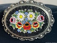 Close-up of oval floral mosaic inlaid with bright colorful flowers on a black background, surrounded by sterling silver frame with scalloped edges and bead accents.
