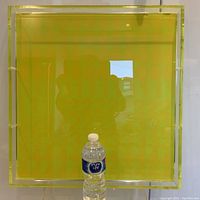 Full frontal view of square acrylic wall art with clear frame and yellow-lime green panel, water bottle for scale