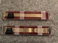 Two gold-tone metal watch bands sealed in original Apollo Flex packaging, one expandable stretch and one link bracelet style.