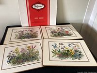Full set of four Pimpernel table mats displayed with original box. Each has a distinct botanical floral print on cream background with gold and brown border.