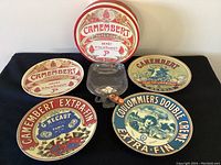 Four round ceramic appetizer plates with vintage cheese label designs arranged on a black cloth. One clear artisan kiln-formed serving dish placed centrally with a cheese spreader on top.