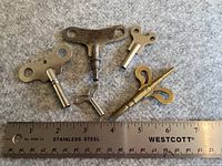 Five vintage clock winding keys placed on a grey fabric with a stainless steel ruler for scale, ranging from about 1.5 to 3 inches in length.