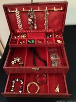 Open wooden jewelry box showing red velvet interior with assorted jewelry organized in compartments including hanging hooks with clip and dangle earrings, and the box lock key placed in bottom drawer.