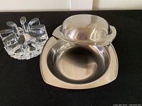 Two stainless steel bowls displayed with the smaller inverted bowl acting as a cover on the larger bowl, next to a heavy, clear glass warner/candle holder.