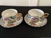 Pair of lustre ware teacup and saucer sets displayed on black background showing overall design, floral motifs, and gold accents.
