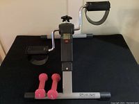 Full view of pedal exerciser on black table with two pink 1-pound weights in front.