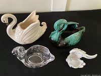 Four swan figurines displayed together: cream ceramic swan planter, green/black ceramic swan, clear lead crystal swan, and small white ceramic swan.