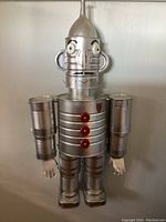 Full view of the Tinman sculpture showing jointed arms, ribbed torso with red buttons, metal hands, and overall silver finish.