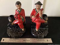 Pair of vintage chalk ware book ends with Asian figures and black glazed jar candle holders. A ruler is placed at their base for scale, showing each figure in red clothing seated on a dark textured base.