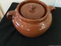 Full view of brown glazed ceramic bean pot with lid and dual side handles on a black surface.