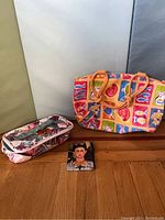 Three assorted bags: colorful large tote bag with food-themed prints, floral patterned travel organizer pouch, and small Frida Kahlo wallet on wooden floor with neutral background.