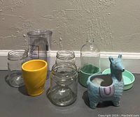 Assorted glass jars, a yellow cup, a blue llama-shaped ceramic planter, and green trays on display surface.
