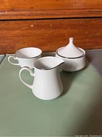 Photo showing two white fine china teacups, creamer, and sugar bowl with silver band trim against wooden background.