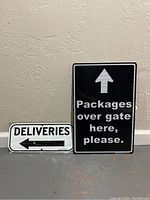 Two metal delivery/package direction signs side by side against a wall, one white with 'Deliveries' and left arrow, the other black with 'Packages over gate here, please.' and upward arrow