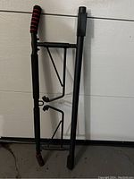 Black metal adjustable pull up bar with foam grips leaning against a white garage door on a concrete floor.