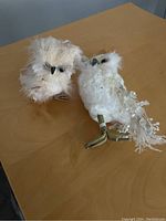 Two decorative owl figurines on a wooden surface, one cream-colored with fluffy feathers and black eyes, the other white with beaded details on wings and tail and greenish metal legs.