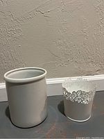 Photo showing two white planter pots: one plain ceramic pot and one decorative metal pot with lace cutouts.