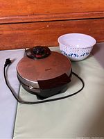Oster stainless steel waffle maker with black handle and temperature control knob, cord included, next to a white mixing bowl with blue horses and patterns.