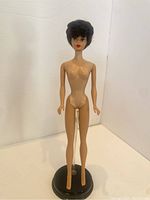 Front view of the nude brunette Bubblecut Barbie doll on a black display stand (not included). The doll has a classic slender Barbie figure and short curly brunette hair.